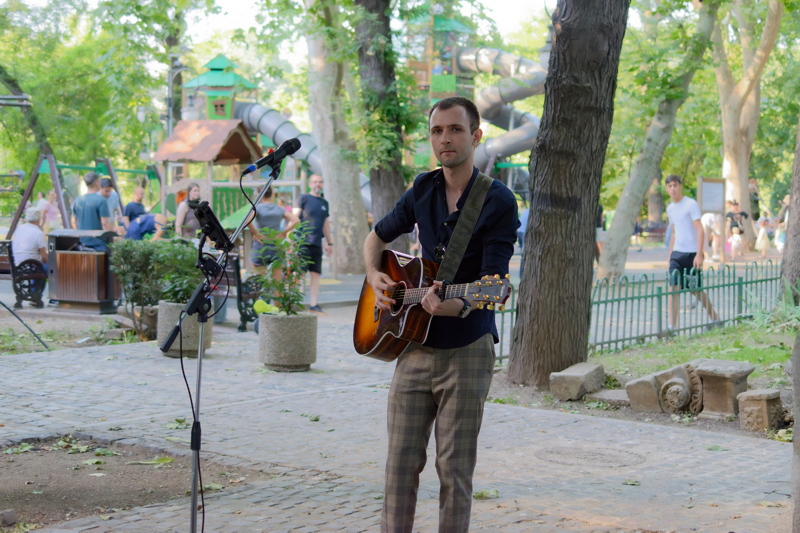 4 - A person playing an acoustic guitar while wearing a black button-down shirt paired with patterned trousers